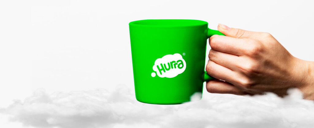 Taza de café con el logo de Hurra, una mano sujetando la tasa. Imagen de fondo blanco, taza color verde. 